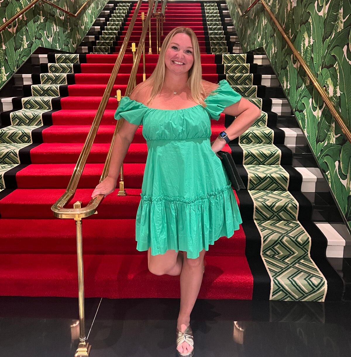 Smiling woman in a green dress posing on a red-carpeted staircase with patterned walls.